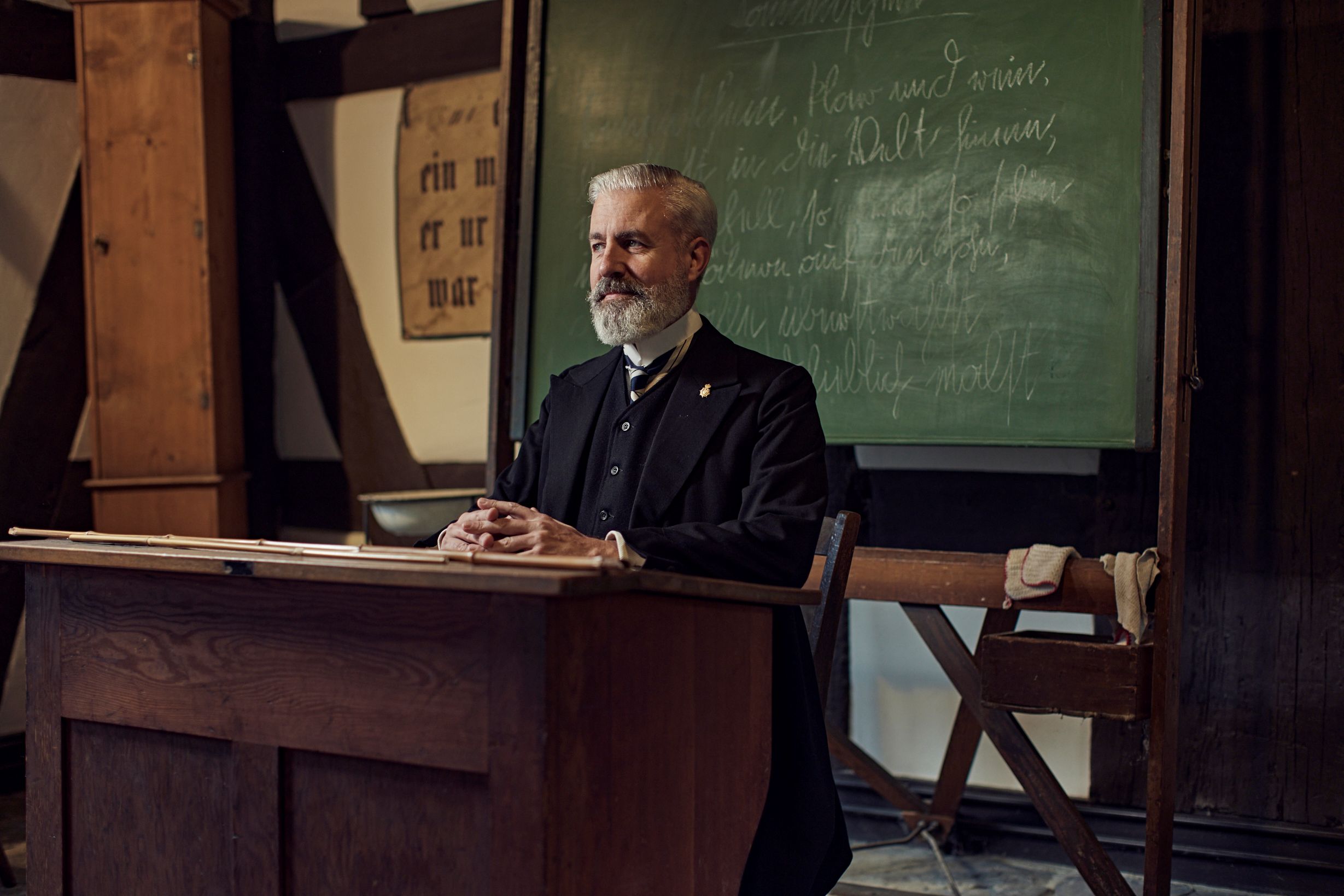 Ein Lehrer im Anzug sitz im Anzug an seinem Pult vor einer Tage, die in Kurrent beschreiben wurde.