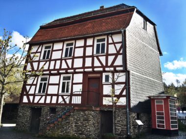 Fackwerkhaus aus Idstein mit Steinsockel und verschieferter Wetterfront, vor der eine alte, roteTelefonzelle steht.